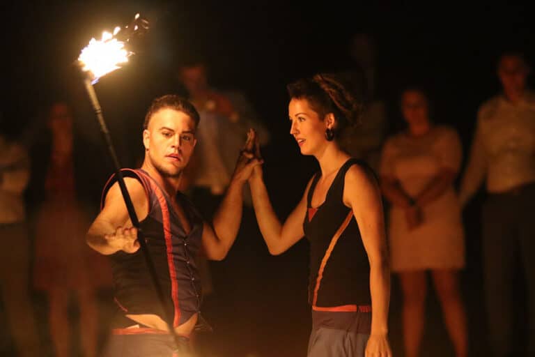 Ein männlicher und eine weibliche Performer stehen nebeneinander, während der Mann eine brennende Fackel in der Hand hält und konzentriert nach vorne schaut. Beide tragen passende, dunkle Bühnenoutfits mit roten Akzenten. Die Frau hält die Hand des Mannes, während sie ihn aufmerksam ansieht. Im Hintergrund steht unscharf eine Gruppe von Zuschauern, die das Geschehen beobachten. Das warme Licht der Fackel beleuchtet ihre Gesichter und verstärkt die intensive und fokussierte Atmosphäre der Szene