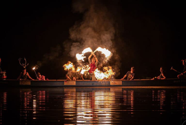 Eine Gruppe von Performern sitzt in einem langen Boot auf einem dunklen Gewässer, während der Hauptakteur in der Mitte des Bootes mit brennenden Fackeln eine Feuershow vorführt. Flammen steigen in die Luft, während der Künstler in einem roten Outfit seine Arme kreuzt und die Fackeln schwingt. Die Flammen und deren Reflexionen auf dem Wasser erzeugen eine dramatische und mystische Atmosphäre. Weitere Personen im Boot, teils mit Requisiten wie Hirschgeweihen, beobachten die Szene. Der Hintergrund ist in tiefes Dunkel gehüllt, was die Feuershow besonders hervorhebt