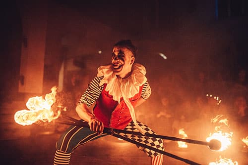 Ein Performer in einem bunten Clownskostüm, bestehend aus einer rot-schwarzen Weste, gestreiften Ärmeln und karierten Hosen, jongliert mit einem brennenden Stab. Er trägt eine große, weiße Rüsche um den Hals und hat dramatisches Gesichtsmakeup. Um ihn herum lodern Flammen, die die Szene in ein warmes, intensives Licht tauchen. Der Performer lächelt breit, während er in einer energiegeladenen Pose steht, umgeben von feurigen Elementen und einer dunklen, atmosphärischen Umgebung.