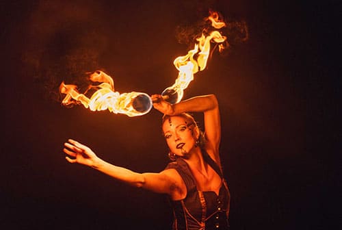 Eine Artistin mit elegantem Makeup und einem dunklen Bühnenoutfit schwingt eine brennende Fackel in einer fließenden Bewegung. Die Flammen tanzen um die Fackel herum und erleuchten die Szene, während die Performerin konzentriert in die Ferne blickt. Die dunkle Umgebung hebt das feurige Licht und die dynamische Bewegung besonders hervor.